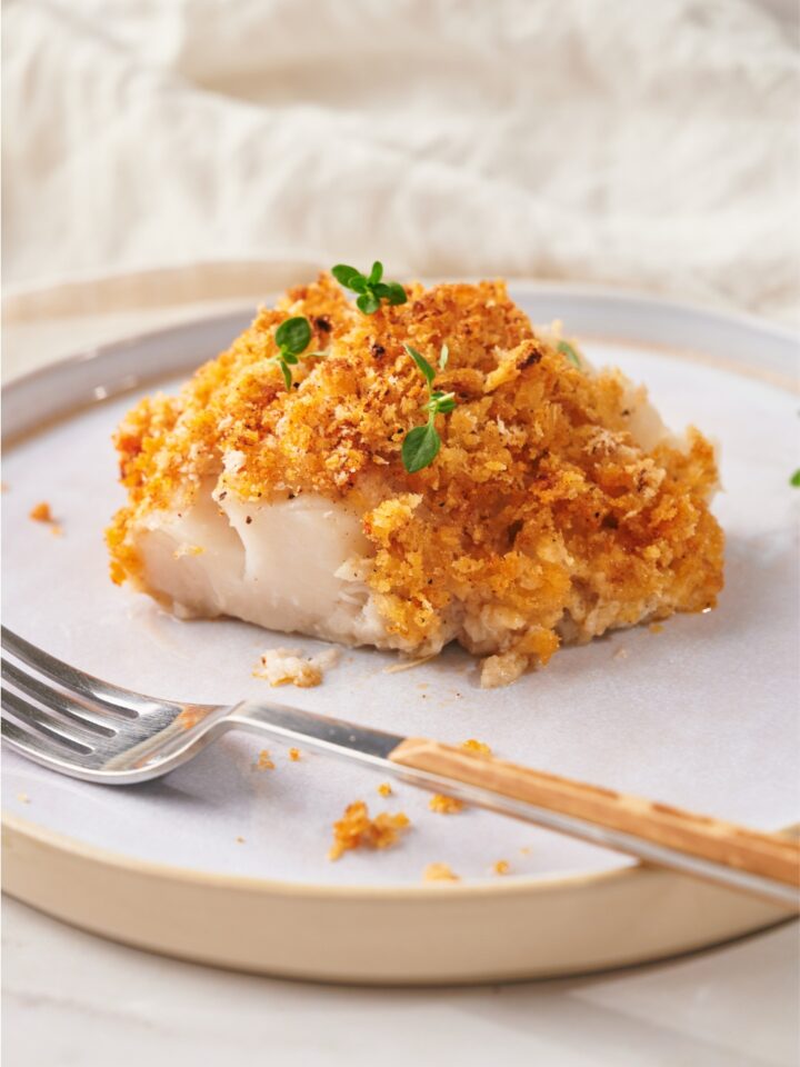 A cod fillet topped with a golden brown bread crumb crust and garnished with fresh herbs. There is a fork on the plate.