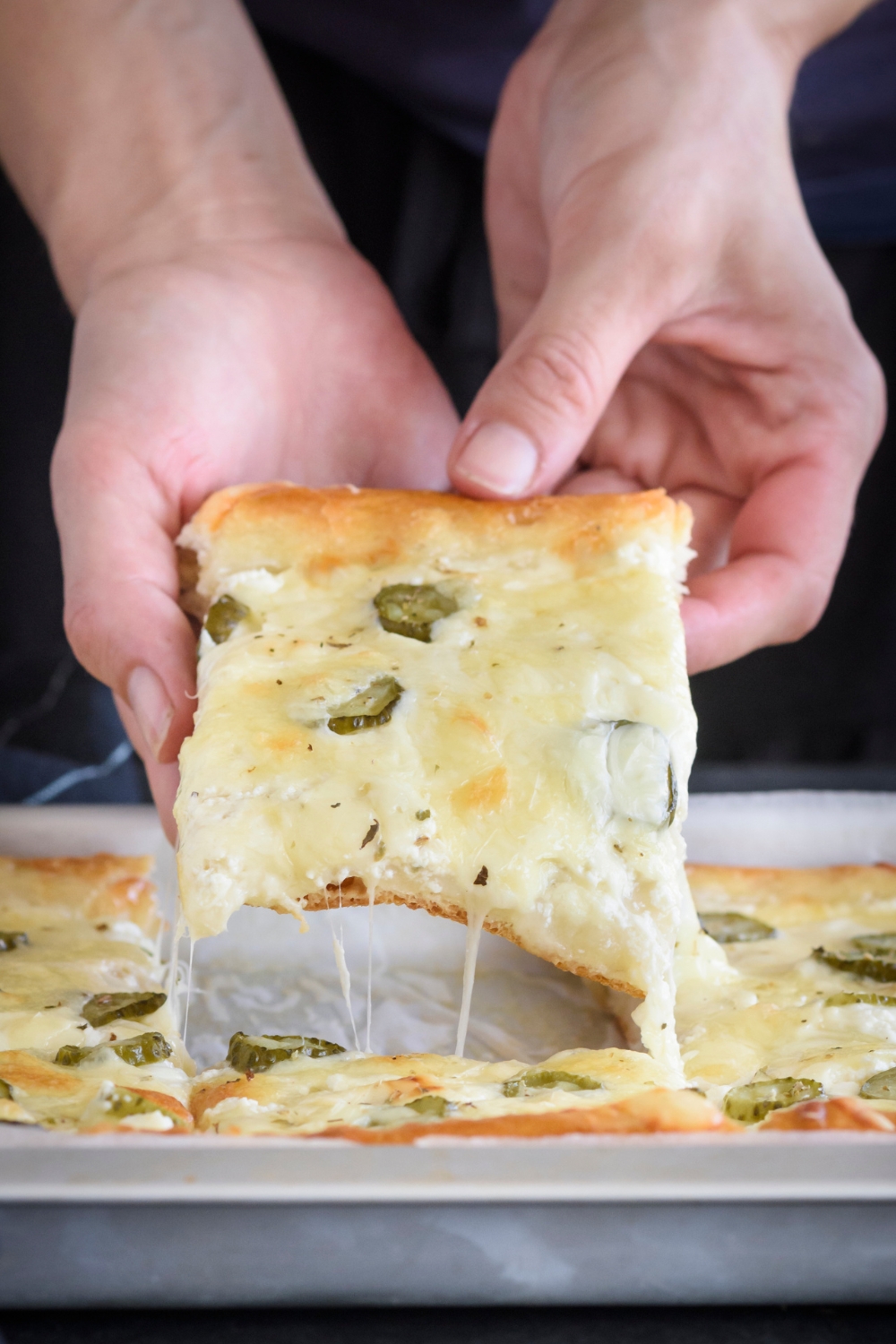 A person removing a slice of pickle pizza from a baking sheet with stringy cheese stuck to the slice.