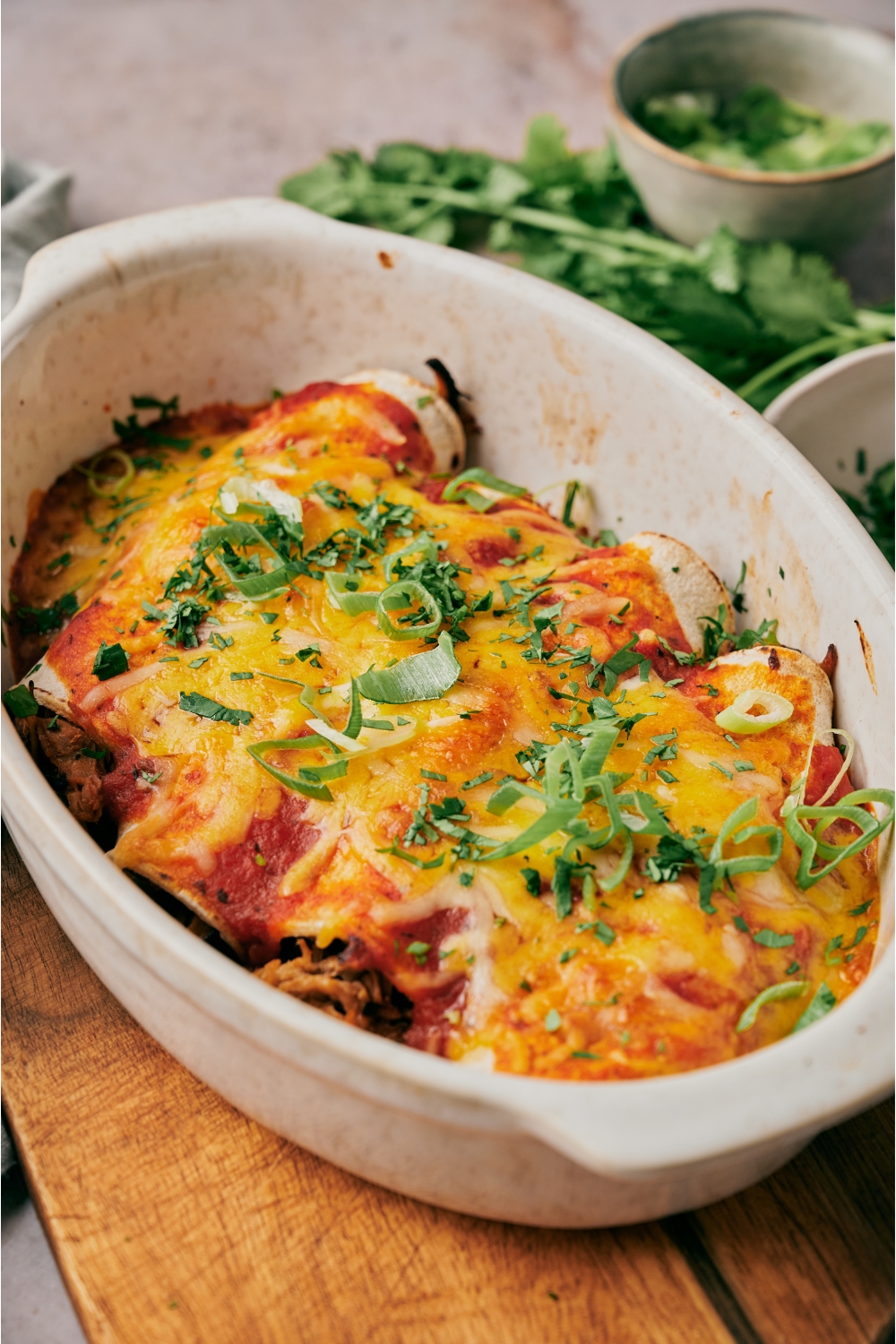 A baking dish atop a wood board filled with freshly baked pork enchiladas covered in melted cheese and fresh herbs.