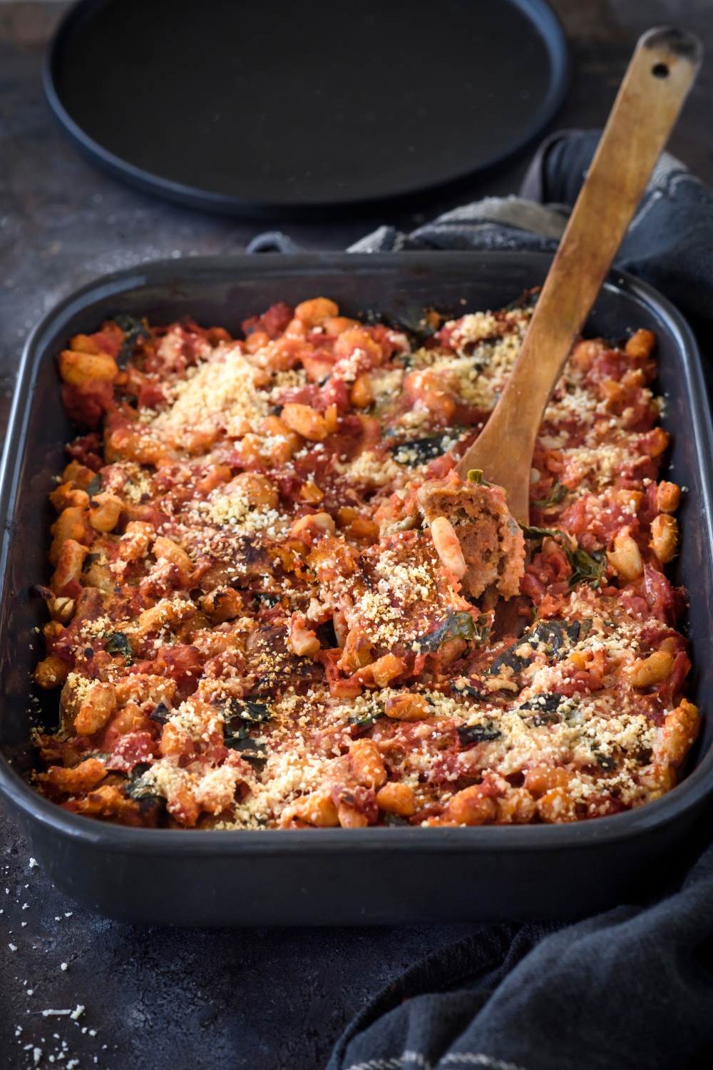 A baking dish filled with tomato casserole covered in parmesan cheese with a wood spoon sticking out of the baking dish.
