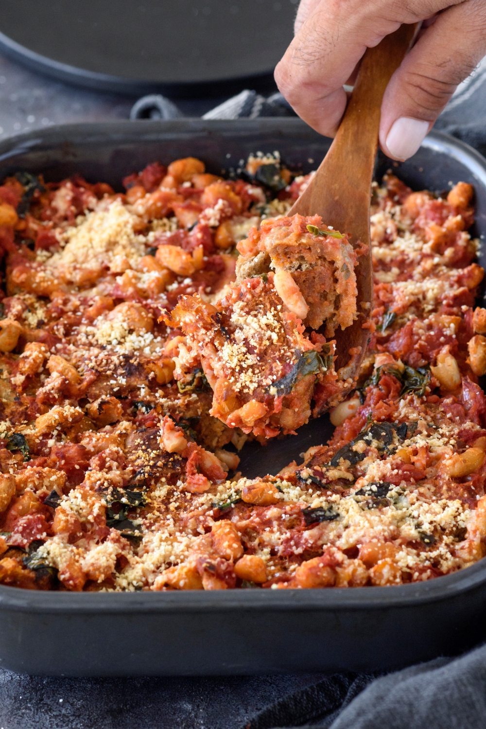 A hand scooping a serving of tomato casserole out of the baking dish using a wooden spoon.