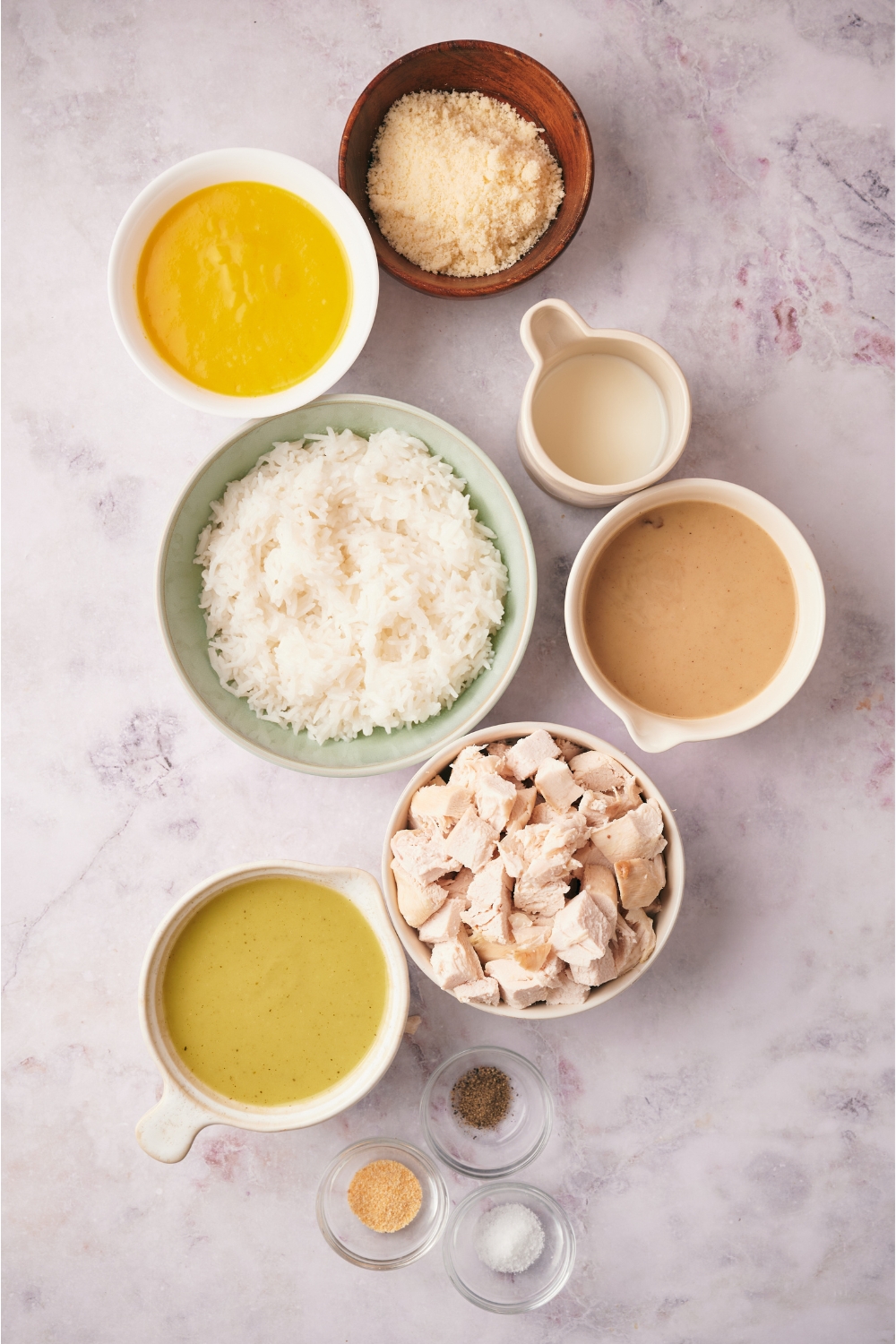 An assortment of ingredients including bowls of cooked white rice, cooked chicken, condensed soup, milk, parmesan cheese, and seasonings.