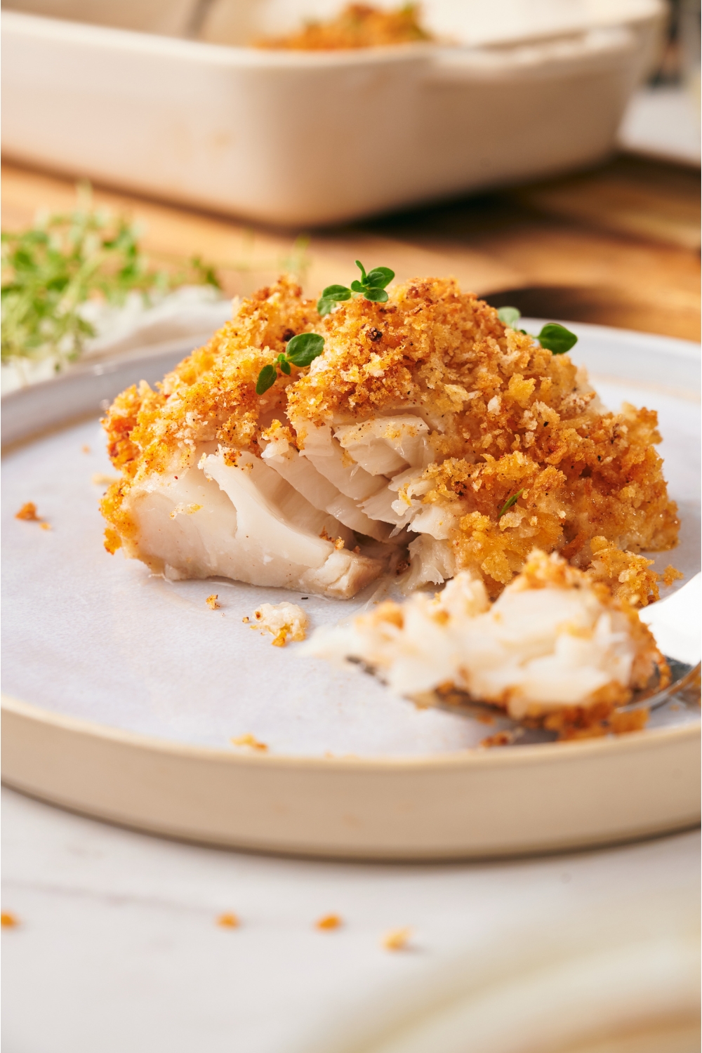 A baked cod fillet topped with golden brown bread crumbs and garnished with fresh herbs. A bite has been taken out and is on a fork on the plate.