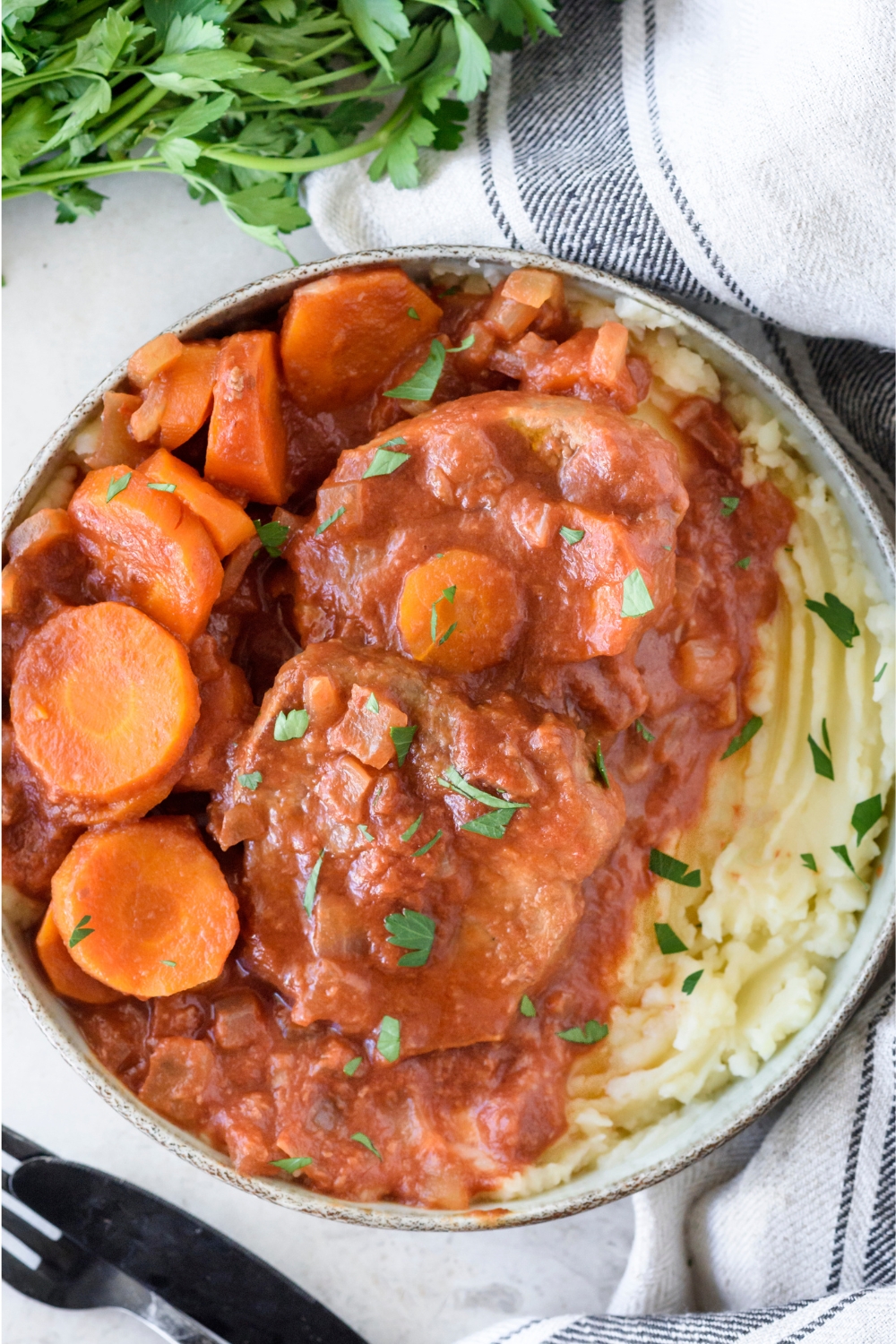 A plate filled with slow cooker swiss steak covered in a red gravy with sides of sliced carrots and mashed potatoes, all covered in a garnish of fresh herbs.