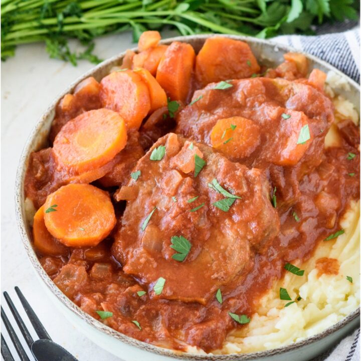 A plate filled with slow cooker swiss steak covered in a red gravy with sides of sliced carrots and mashed potatoes, all covered in a garnish of fresh herbs.