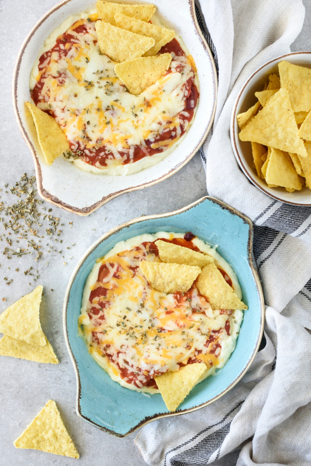 Two baking dishes filled with chili cheese dip covered in a layer of melted cheese with corn chips sticking out of the dip. There is a bowl of corn chips and some corn chips have fallen onto the counter.