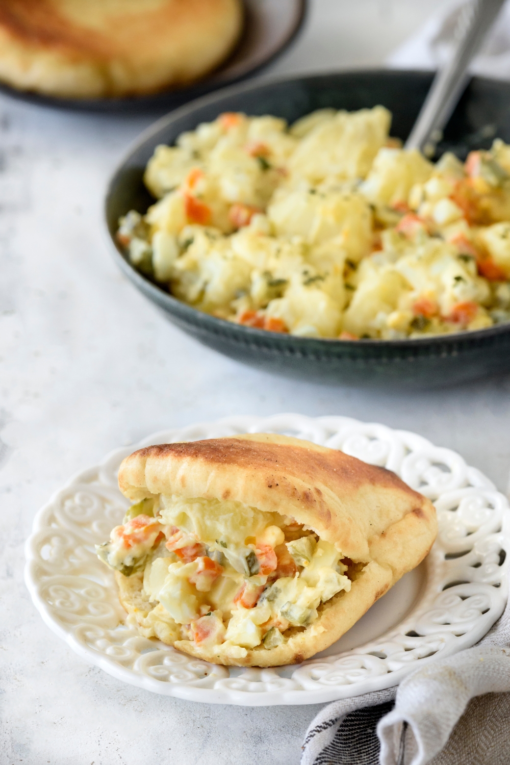 Potato salad stuffed in a pita on a decorative white plate with the rest of the potato salad in the background.