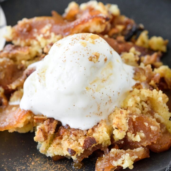 A bowl of apple dump cake with a scoop of vanilla ice cream and a dusting of cinnamon on top. The ice cream is beginning to melt.