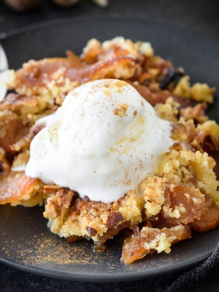 A bowl of apple dump cake with a scoop of vanilla ice cream and a dusting of cinnamon on top. The ice cream is beginning to melt.