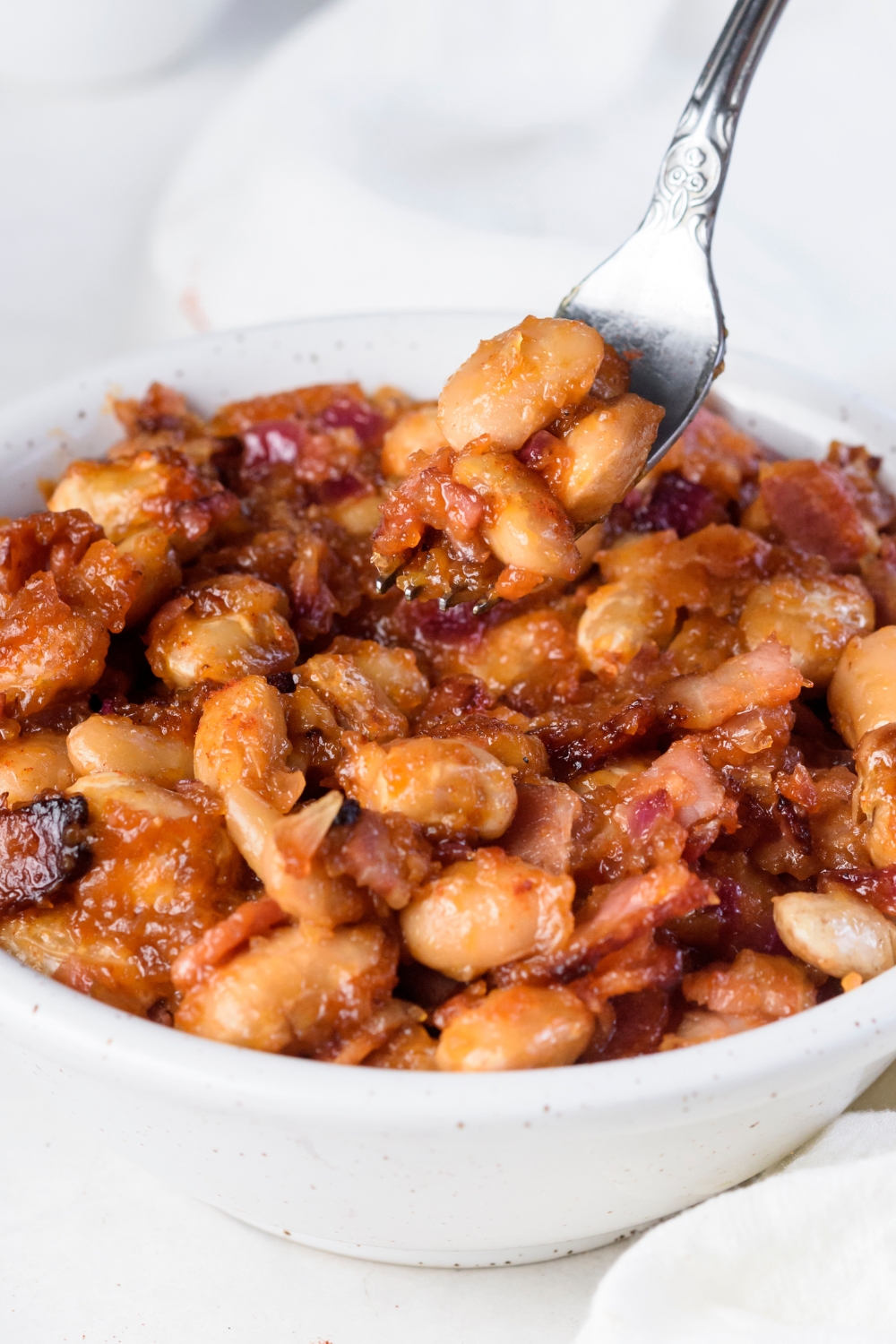 A fork holding a bite of baked beans with crispy bacon coated in sauce. The fork is above a bowl filled with baked beans.