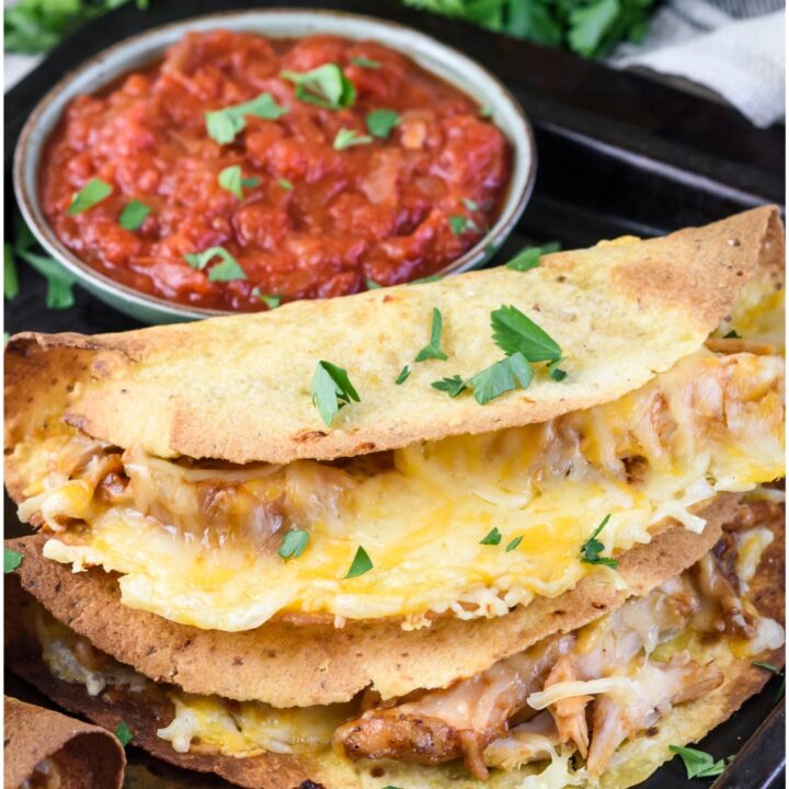 Two crispy tacos stacked on top of each other on a tray with bowls of red and green salsa. Both tacos are filled with cooked and shredded chicken and melted cheese.
