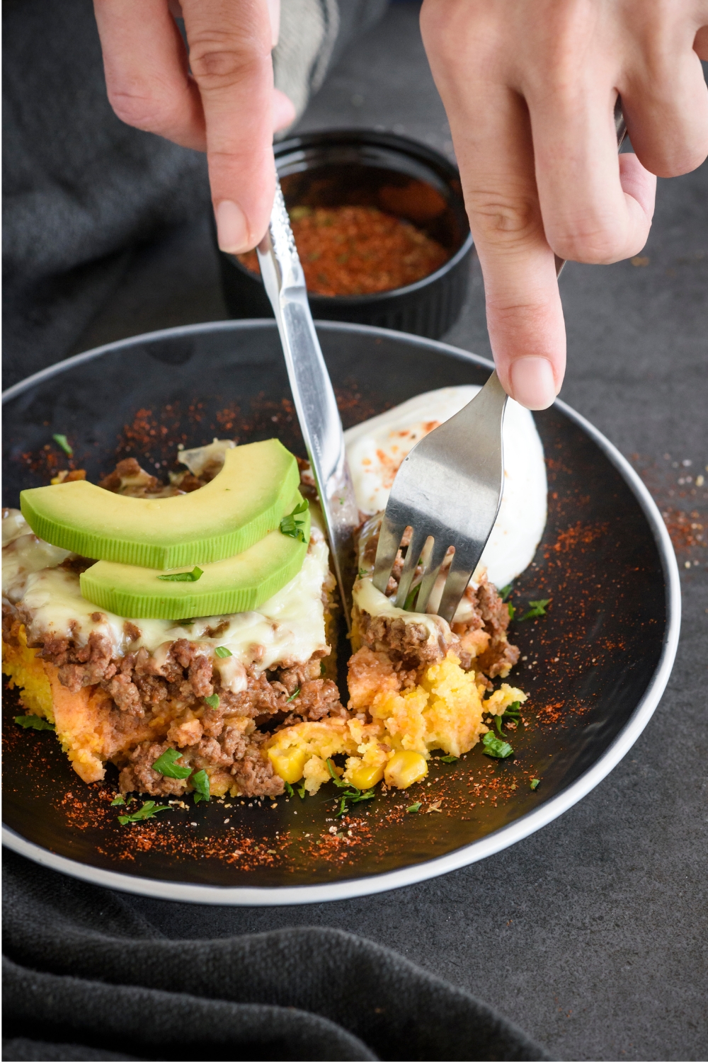 A person using a fork and knife to cut into a serving of beef tamale casserole. The casserole is covered in melted cheese and topped with sour cream and sliced avocados.