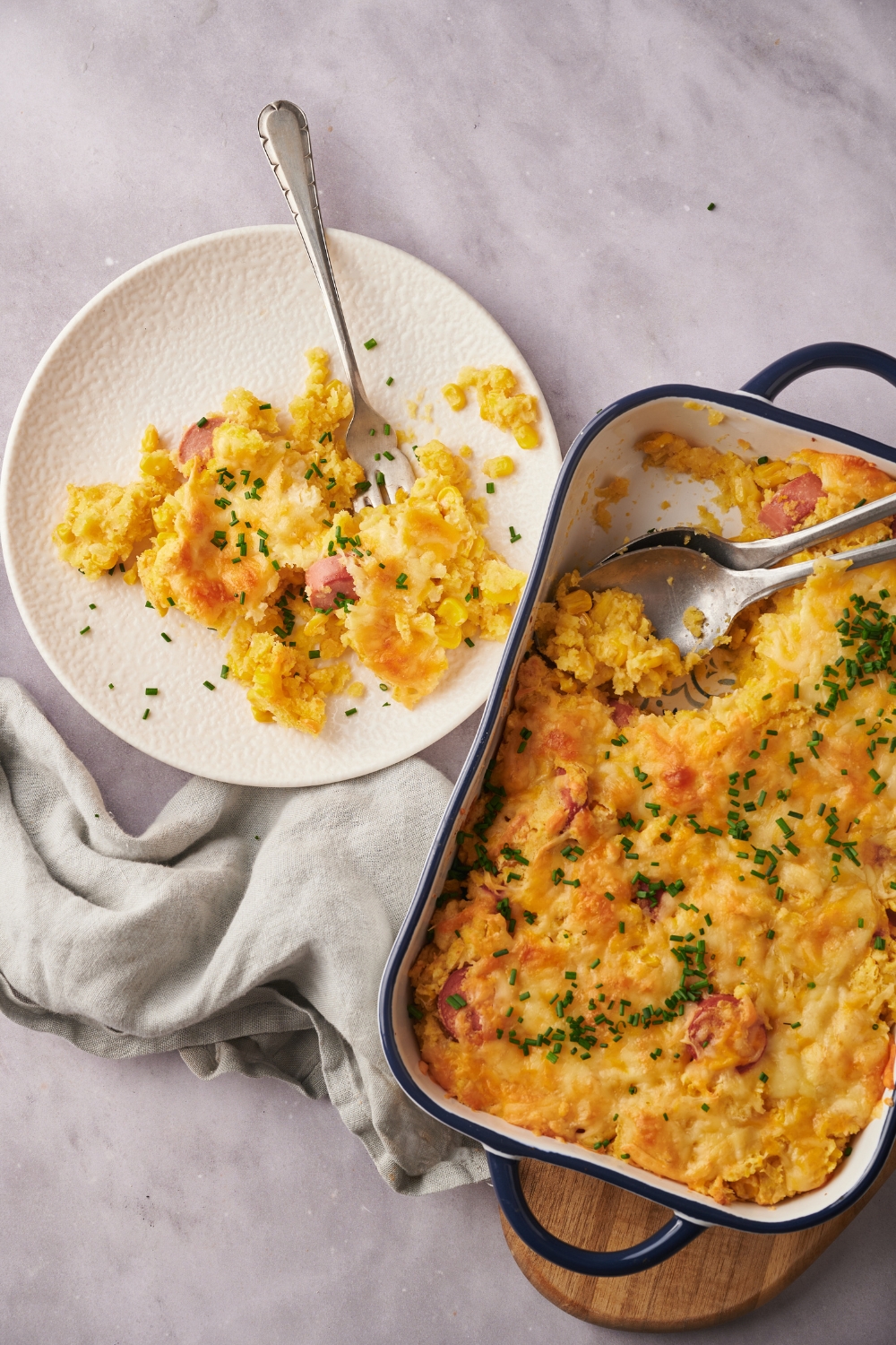 A serving of corn dog casserole next to a baking dish filled with casserole. There is a fork on the plate and the casserole is garnished with chives.