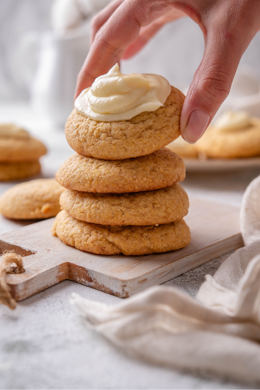 A hand grabbing a cornbread cookie from a stack of four cookies on a wooden board. The top cookie has a dollop of buttercream frosting.