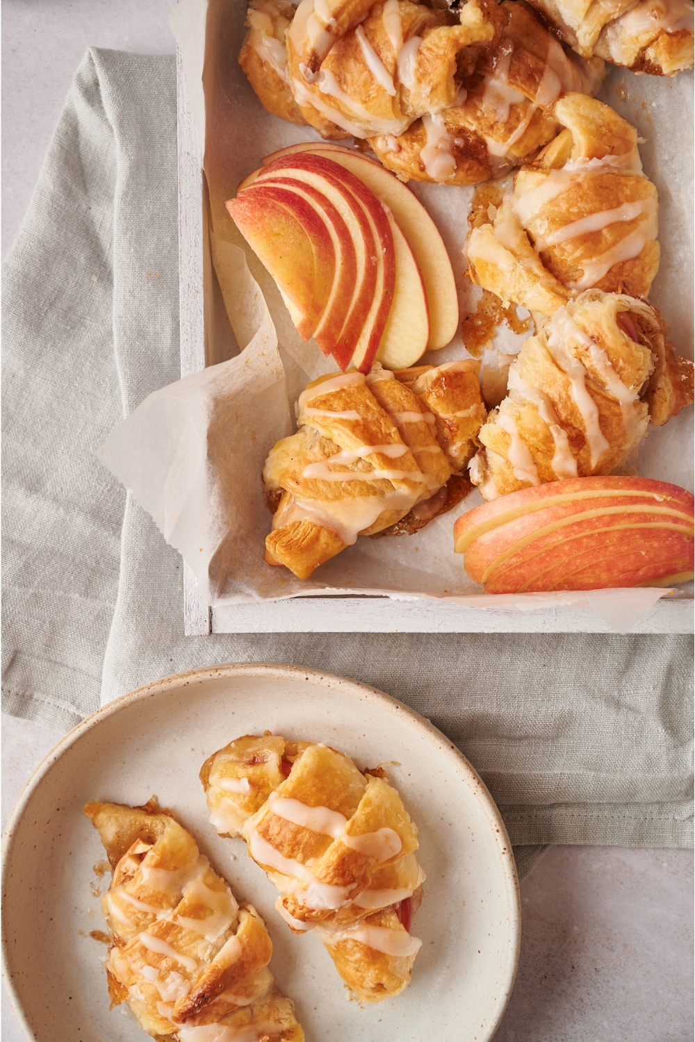 A dish lined with parchment paper filled with baked apple turnovers and apple slices next to a plate with two apple turnovers.