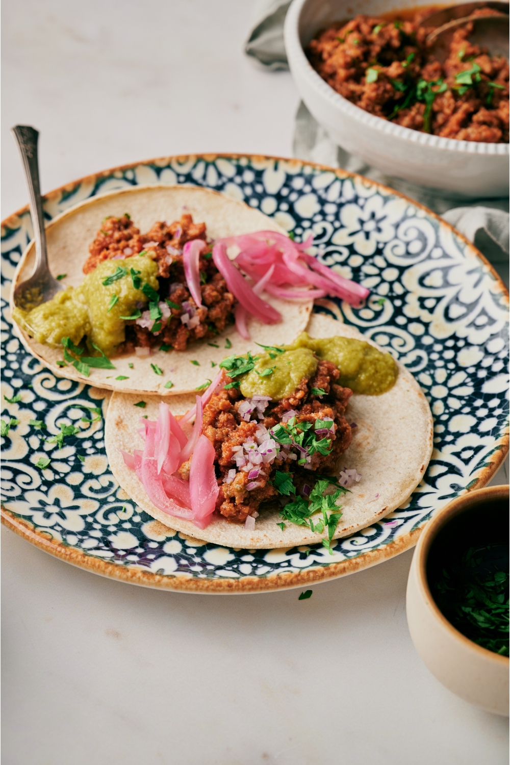 Two tacos on a decorative blue plate topped with taco meat, pickled red onions, fresh herbs, and green salsa.