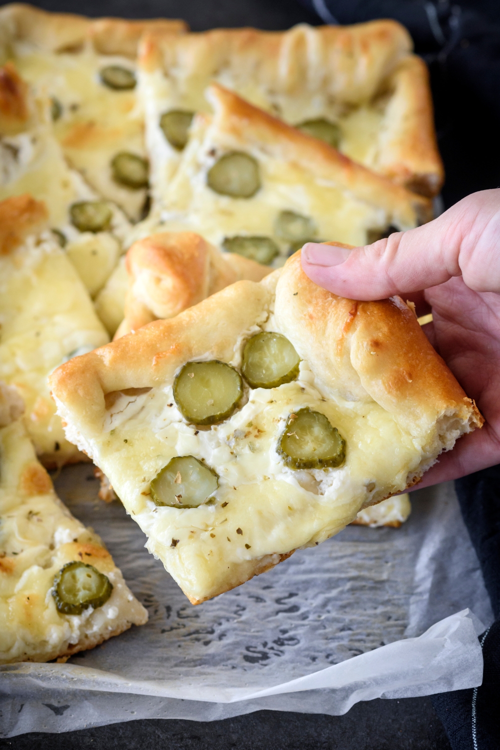A hand holding a square piece of pickle pizza with the rest of the pizza in the background on a sheet of parchment paper.