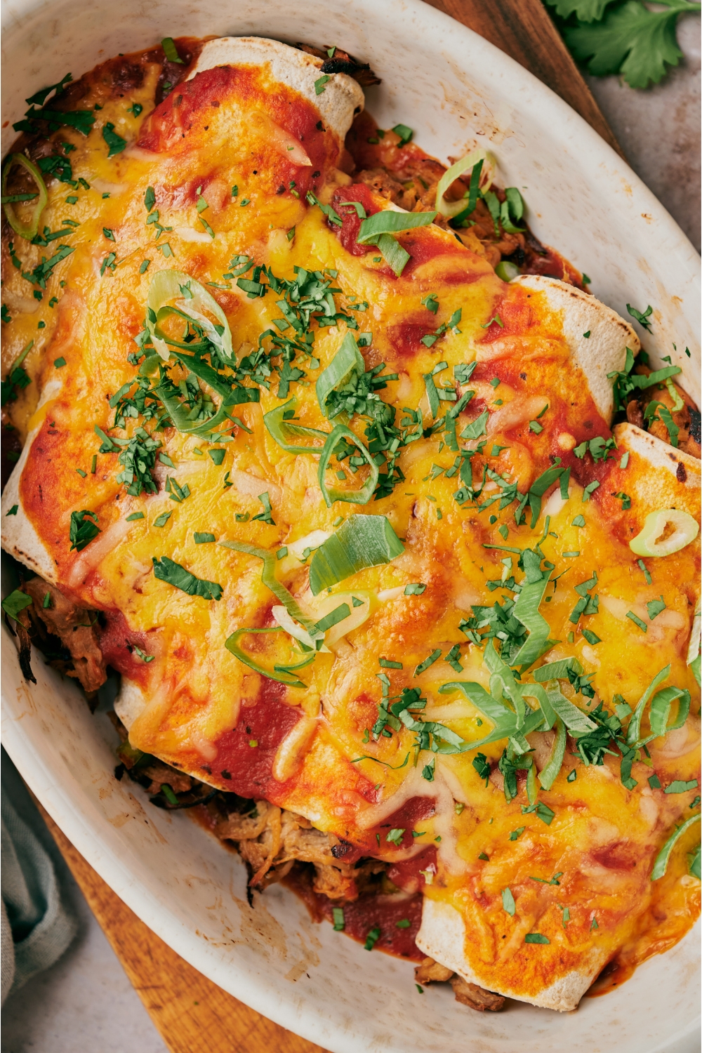 A baking dish atop a wood board filled with freshly baked pork enchiladas covered in melted cheese and fresh herbs.