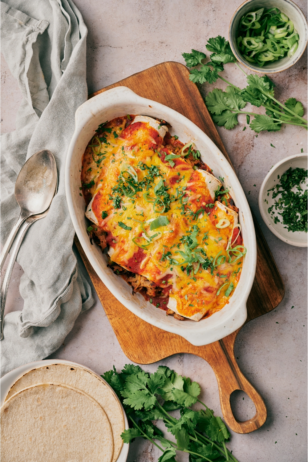 A baking dish filled with freshly baked pork enchiladas covered in melted cheese and fresh green herbs. The baking dish is on a wood board surrounded by fresh cilantro.