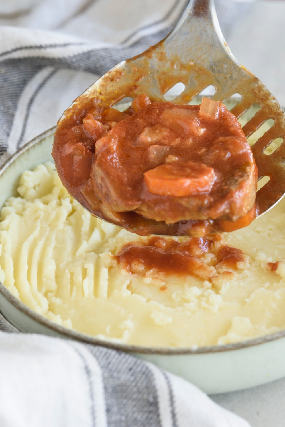 A plate of mashed potatoes with a slotted spoon spooning a cooked swiss steak covered in red gravy with bits of diced onion and carrots mixed with the gravy.