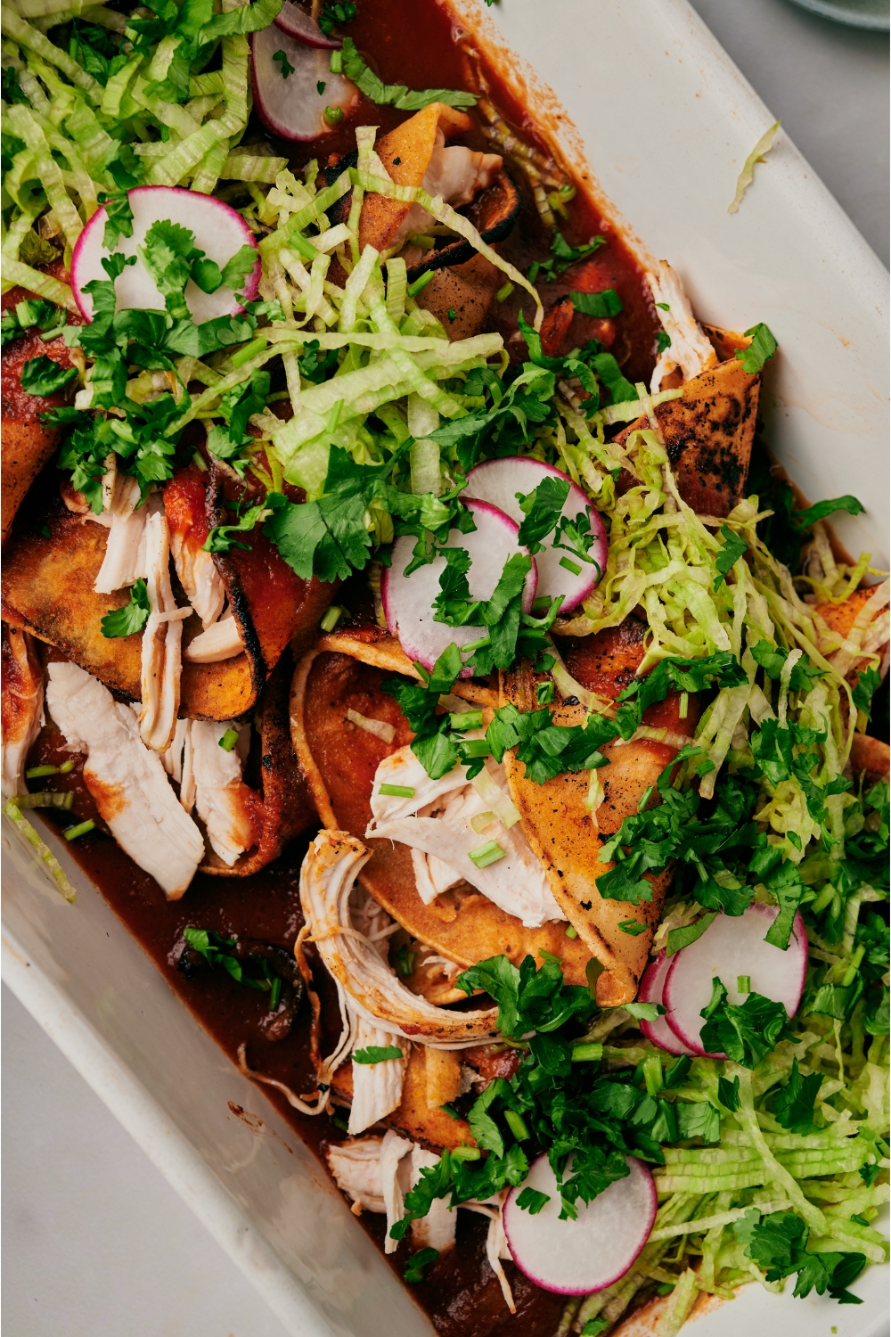 Close up of chicken enchiladas covered in a red chili sauce and garnished with chopped lettuce, cilantro, and sliced radishes.