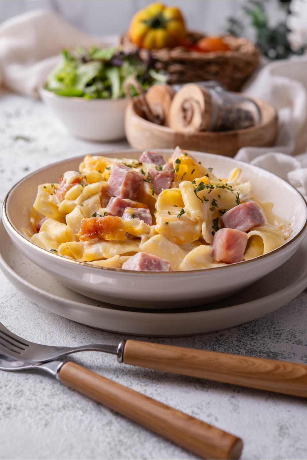 A bowl of ham and noodle casserole garnished with green herbs atop a plate with two forks next to it.