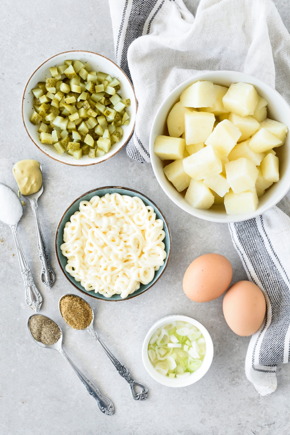 A countertop with diced potatoes, diced pickles, mayo, mustard, seasonings, eggs, and green onions in various bowls.