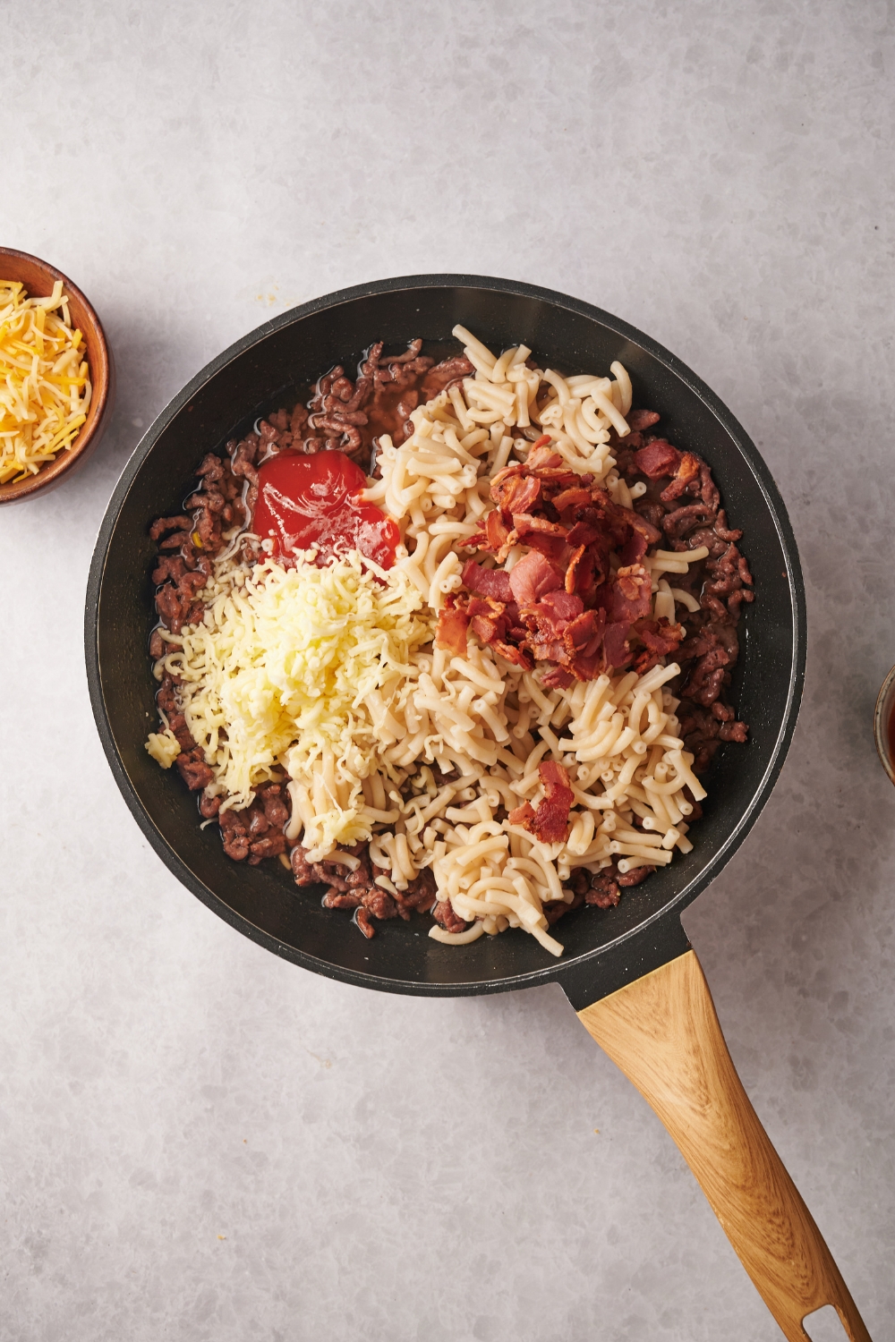 A skillet filled with cooked ground beef, shredded cheese, cooked macaroni noodles, ketchup, and bacon.