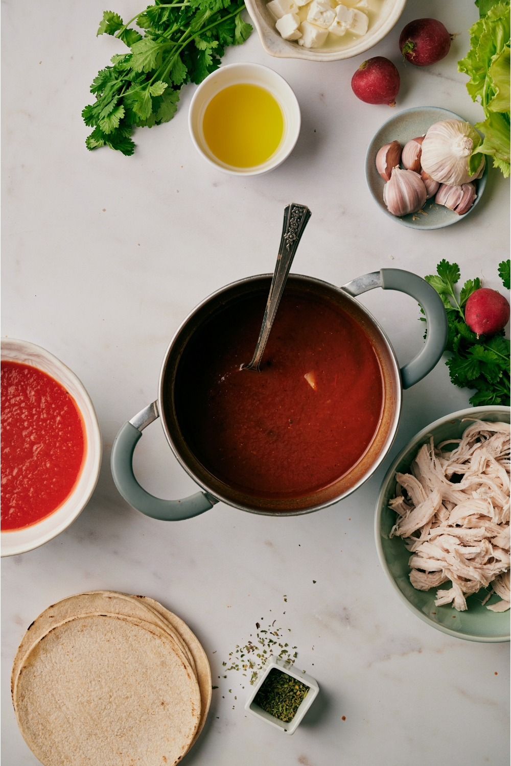 A pot filled with a strained chili sauce mixture. The pot is surrounded by an assortment of ingredients and there is a spoon in the pot.