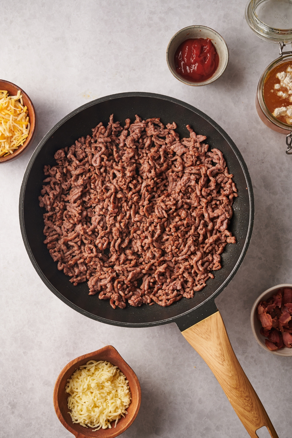 A skillet filled with cooked ground beef. The skillet is surrounded by an assortment of ingredients.