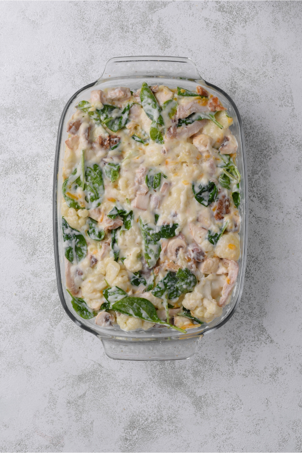 A baking dish filled with unbaked chicken, cauliflower and spinach casserole.