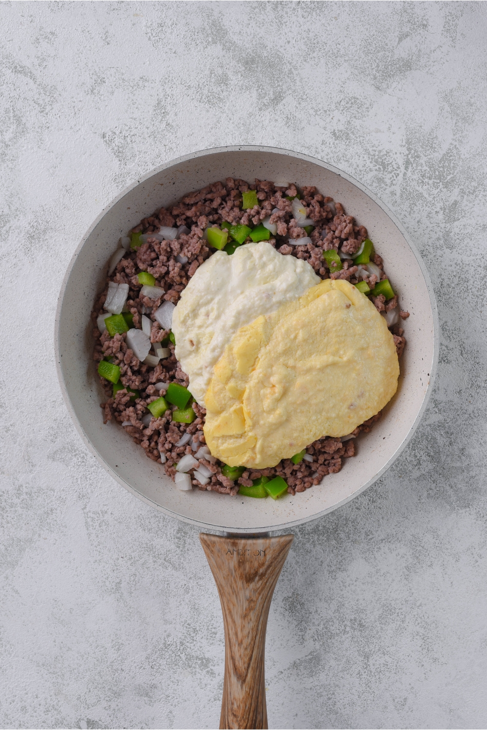 A skillet with cooked ground beef mixed with diced peppers and onions. Two creamy sauces are dumped on top of the beef but not mixed together.