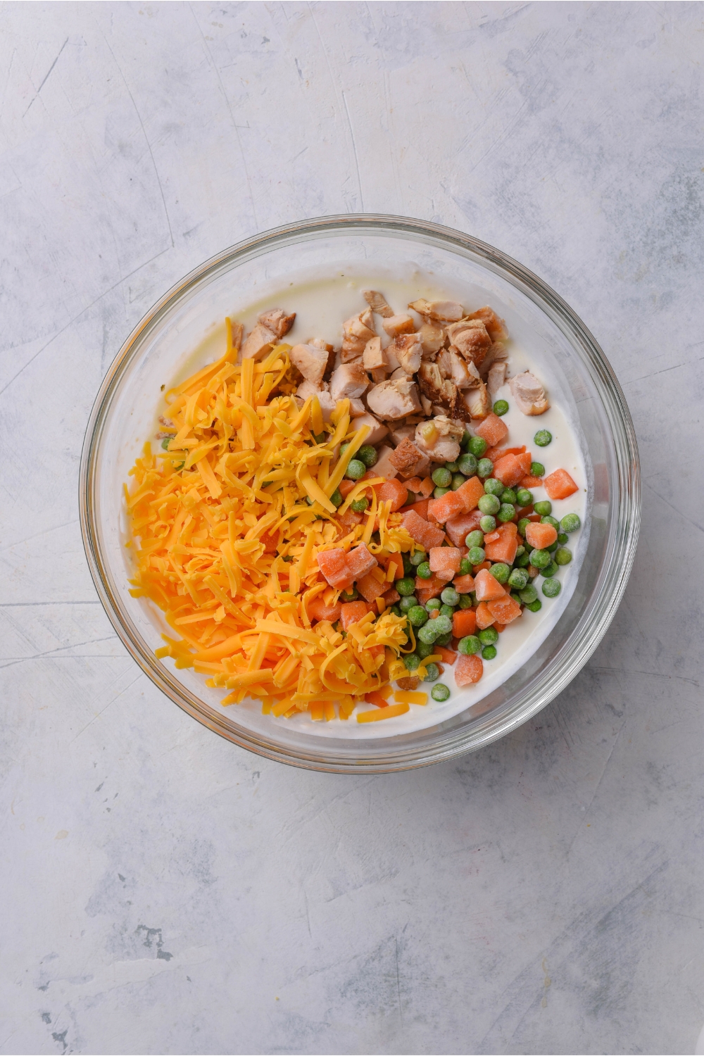 A clear bowl with diced chicken, peas, carrots, shredded cheese, and a creamy soup mixture.