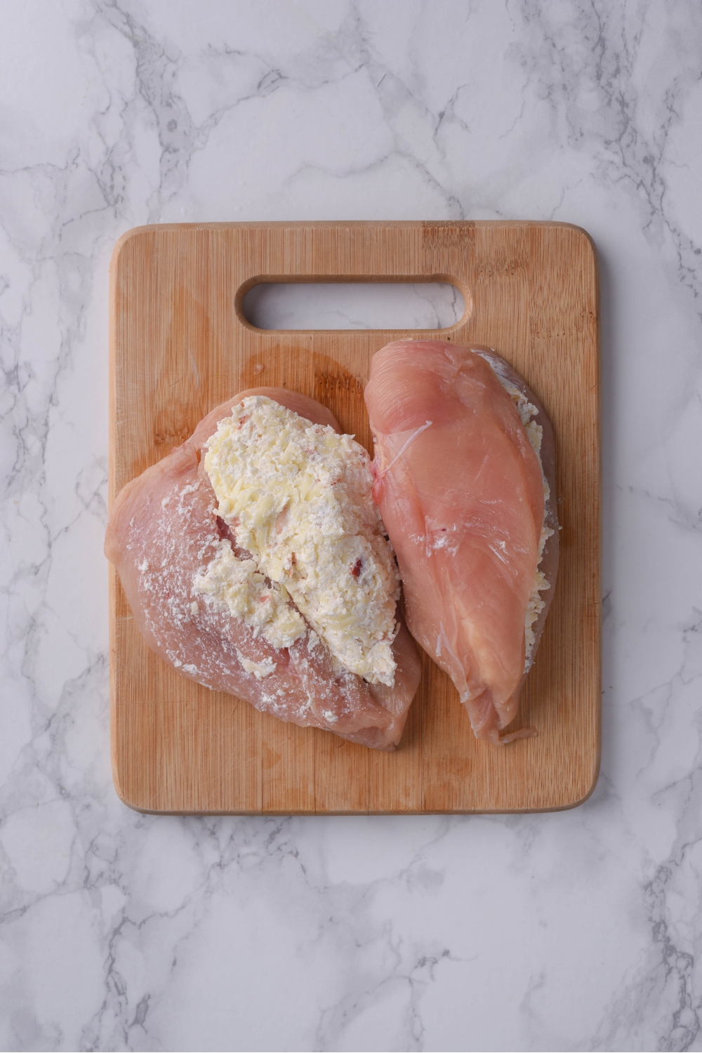 Two raw chicken breasts butterflied on a wood cutting board. Both are stuffed with a cream cheese mixture and one is open.