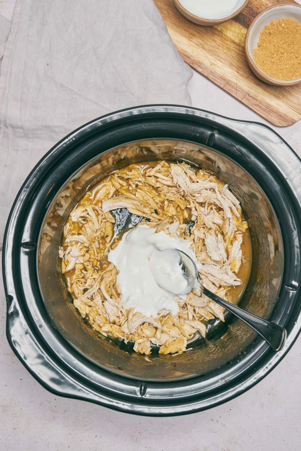 A slow cooker filled with shredded chicken in gravy and a spoonful of sour cream has been added to the slow cooker.