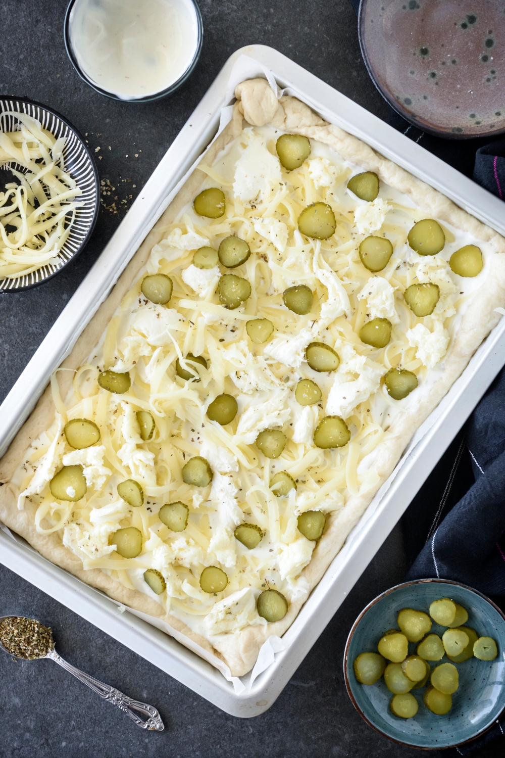 Unbaked pickle pizza on a baking sheet lined with parchment paper. The baking sheet is surrounded by an assortment of ingredients.