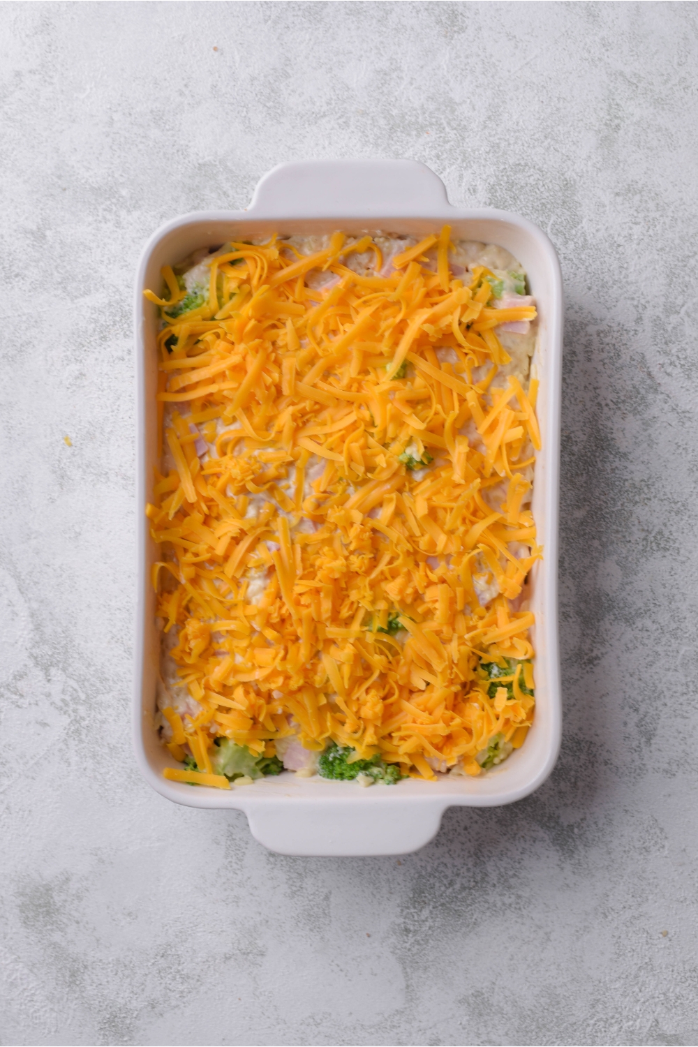A baking dish filled with unbaked ham casserole covered in a layer of shredded cheese.