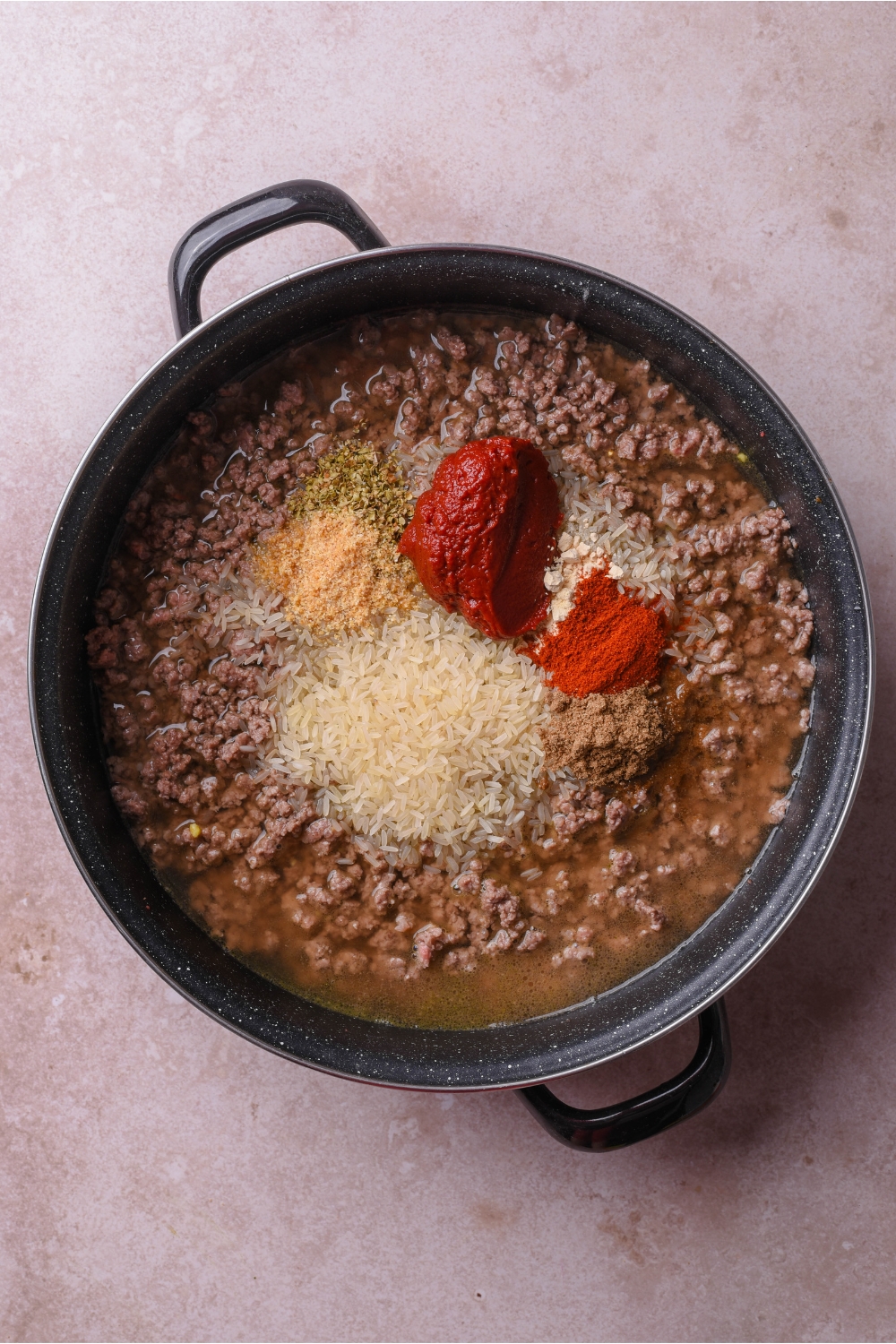 A large skillet with cooked ground beef, broth, tomato pasta, uncooked rice, and an assortment of spices.
