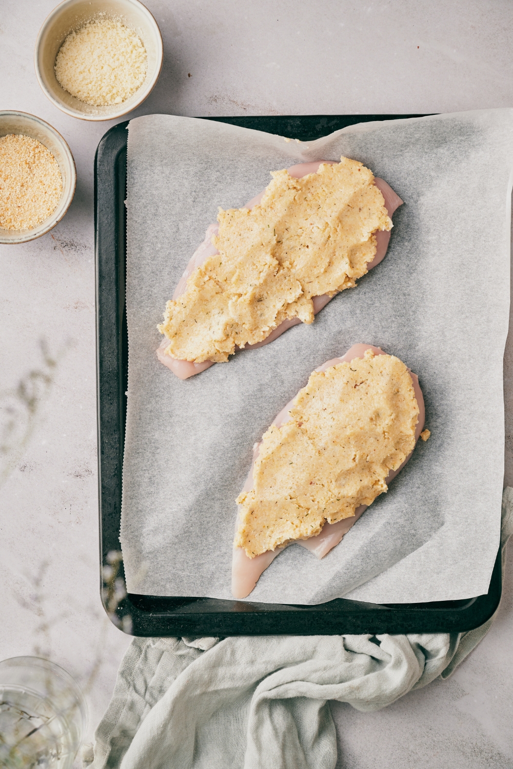 Two raw chicken breasts covered in a seasoned bread crumb mixture on a baking sheet lined with parchment paper.