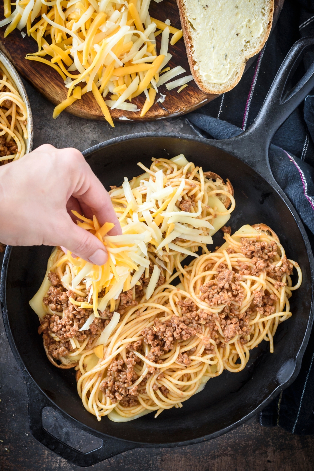 A hand spreading shredded cheese over the top of two spaghetti sandwiches.