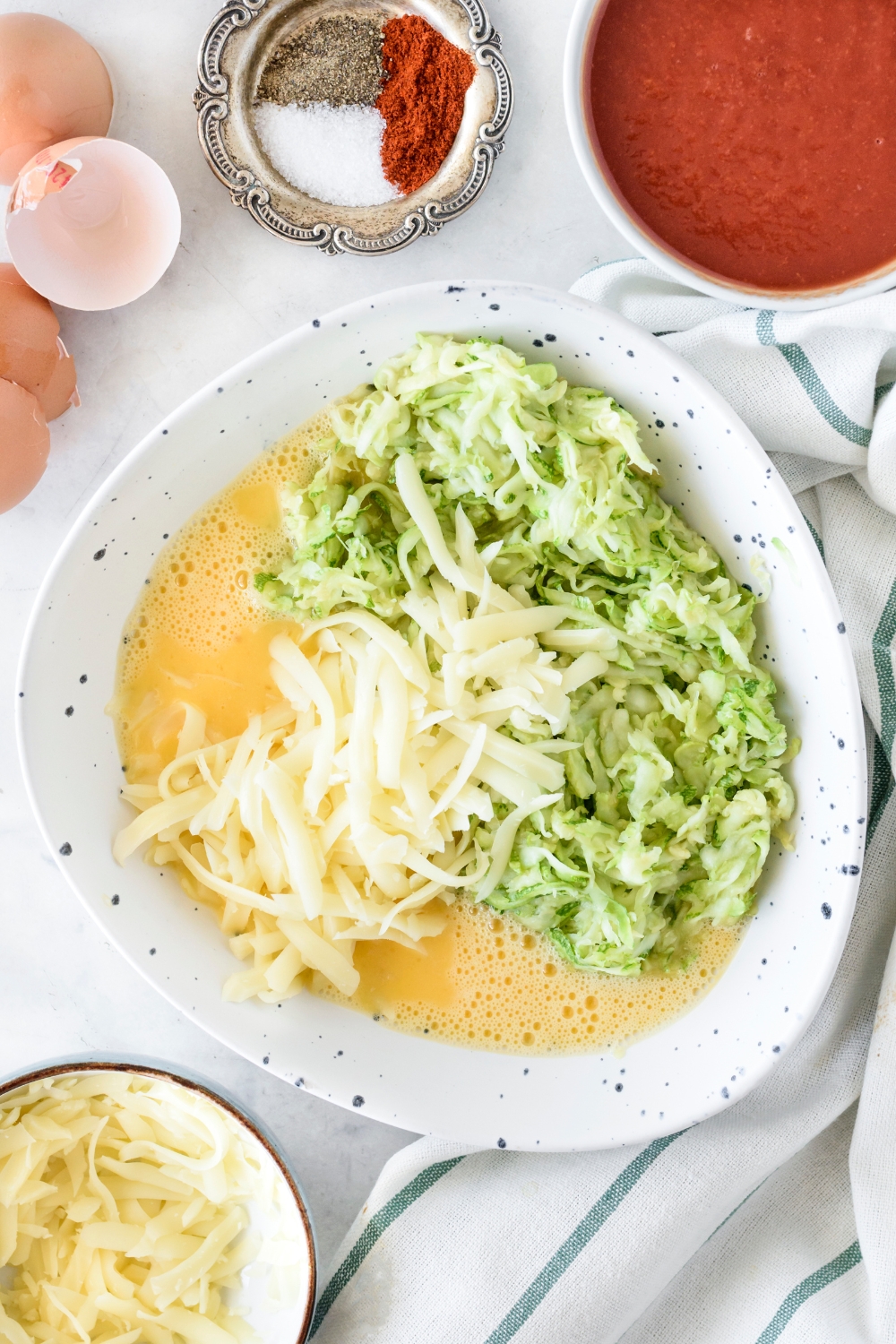 A bowl filled with grated zucchini, beaten eggs, and shredded cheese. The ingredients have not been combined.