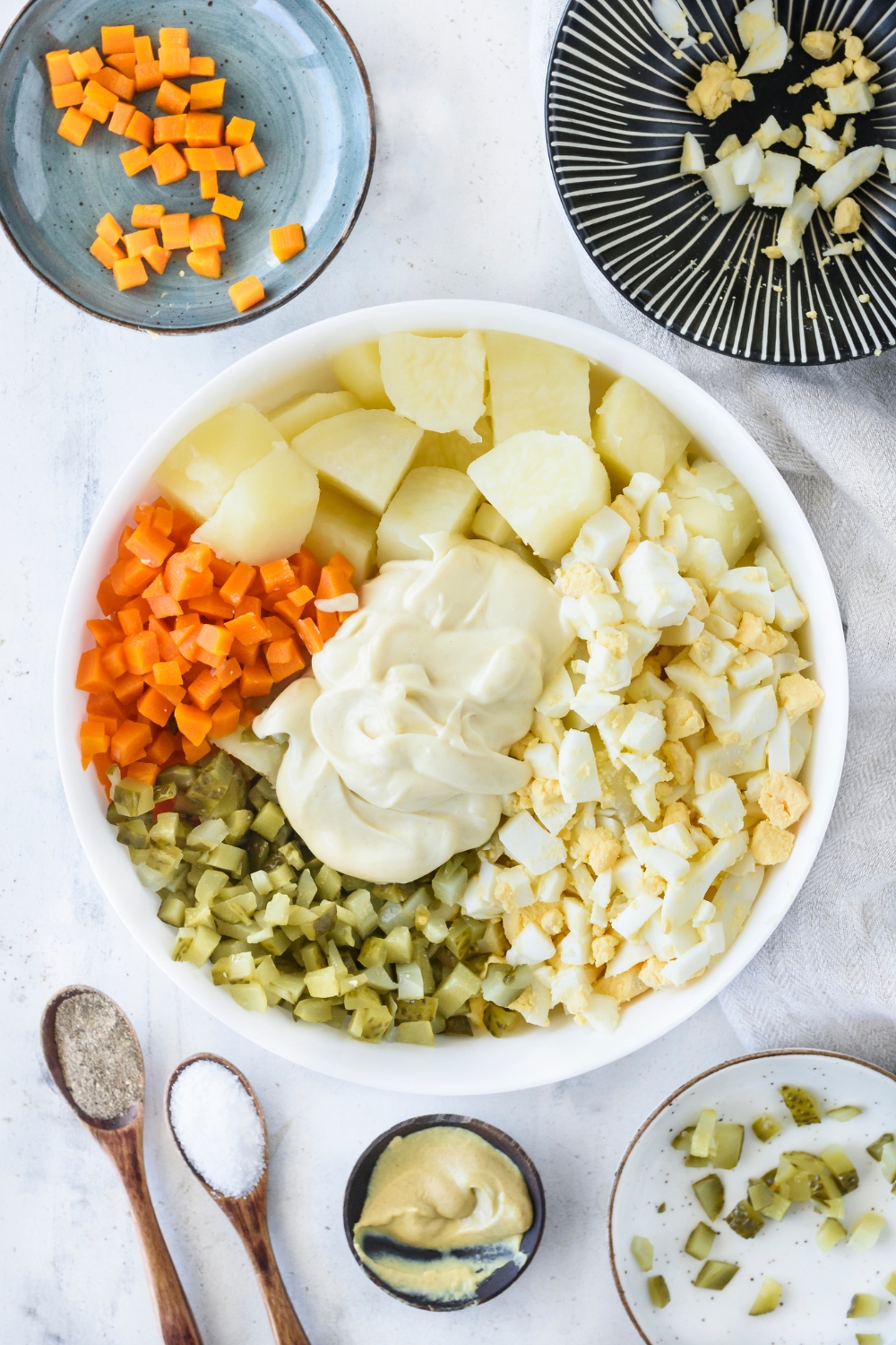 A bowl filled with chopped hard-boiled eggs, diced carrots, diced pickles, boiled potatoes and mayonnaise. The ingredients have not been mixed together.
