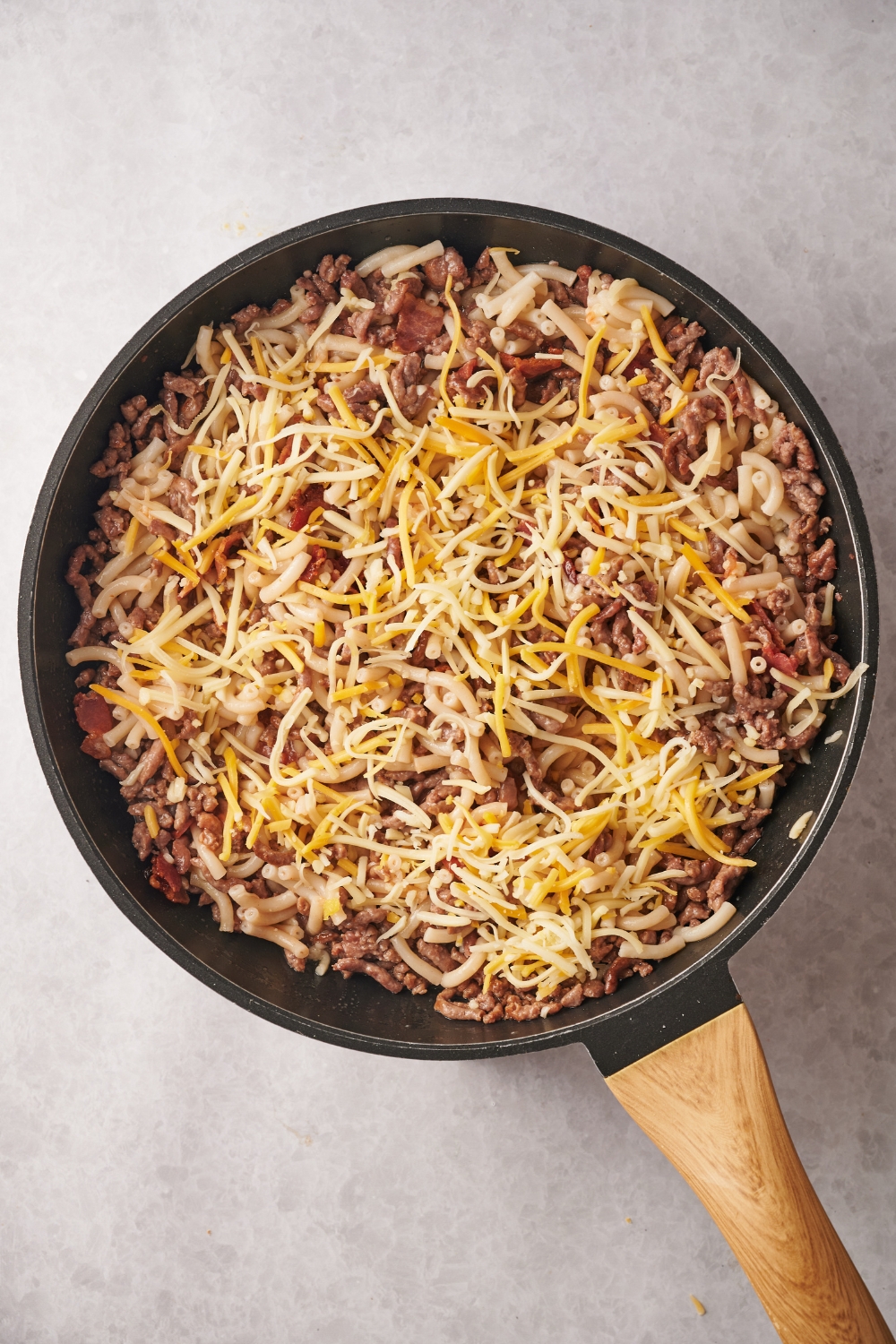 A skillet filled with cooked ground beef, macaroni noodles, and bacon covered in a layer of shredded cheese.