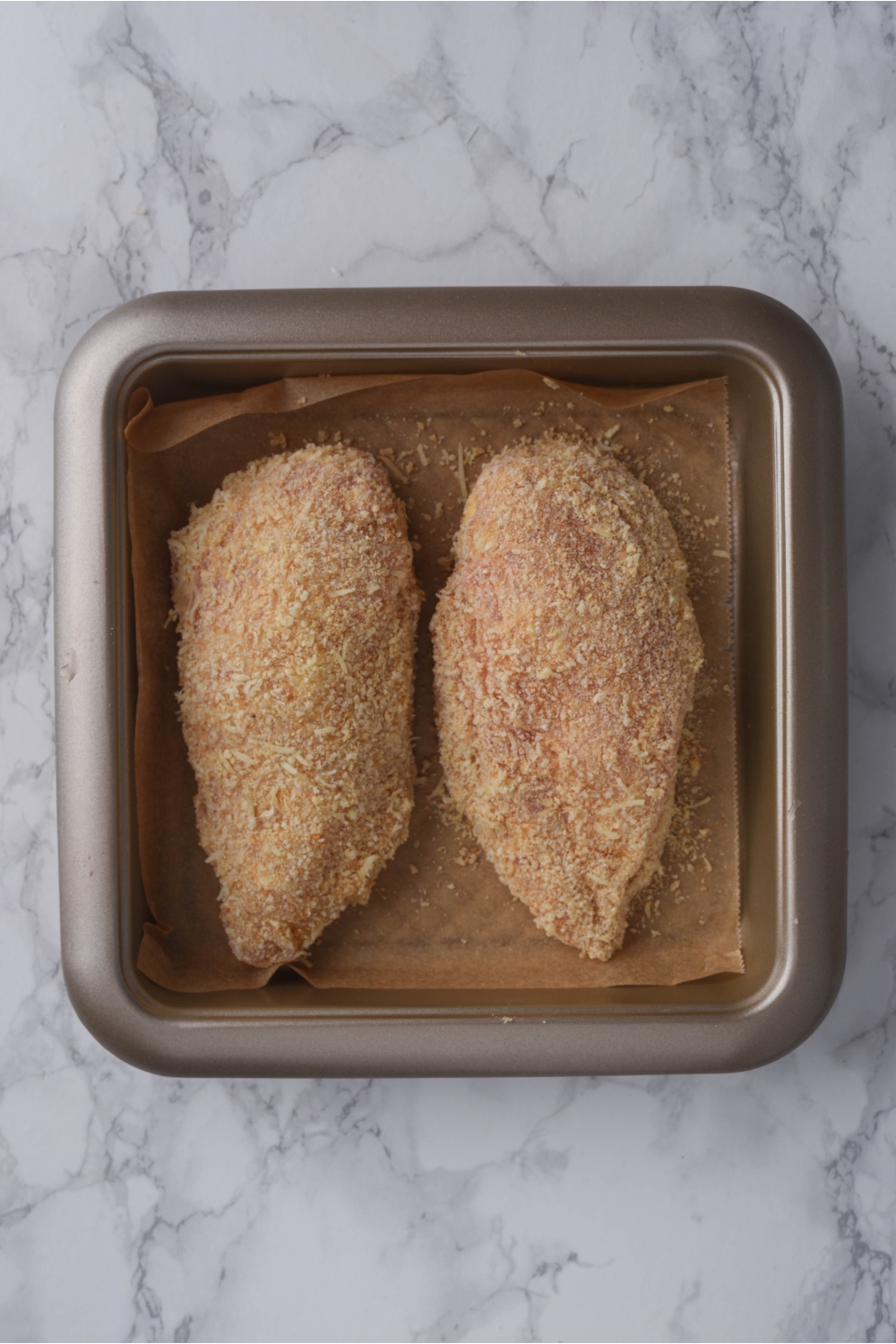 Two raw, breaded chicken breasts in a baking pan lined with parchment paper.