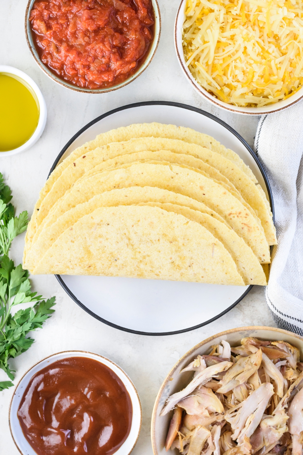 An assortment of ingredients including a plate of corn tortillas and bowls of barbecue sauce, shredded chicken, salsa, shredded cheese, and oil.