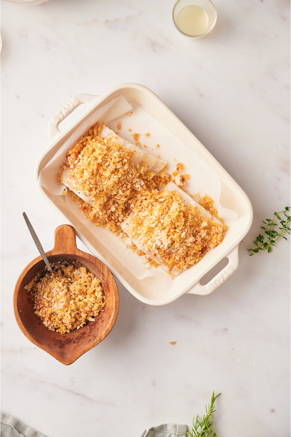 A baking dish with two cod fillets topped with a seasoned bread crumb mixture. The bowl of bread crumbs is next to the baking dish.