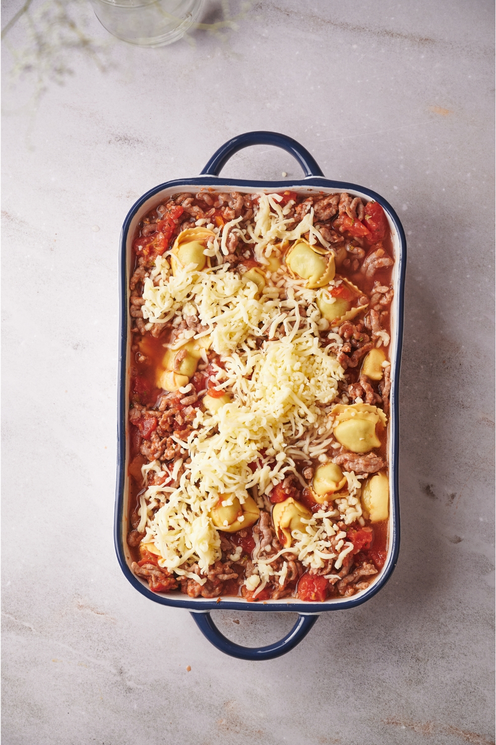 A baking dish filled with cooked ground beef, tomato sauce, tortellini pasta, and shredded cheese on top.
