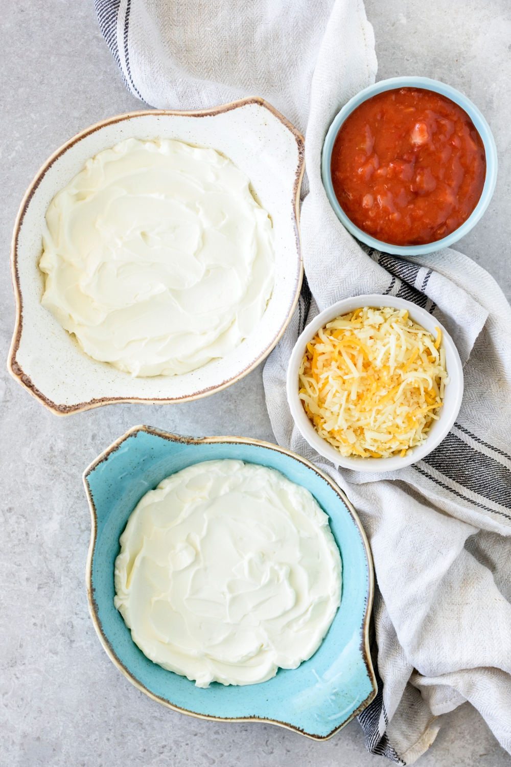 Two baking dishes filled with an even layer of cream cheese. Next to the two baking dishes are bowls of chili and shredded cheese.
