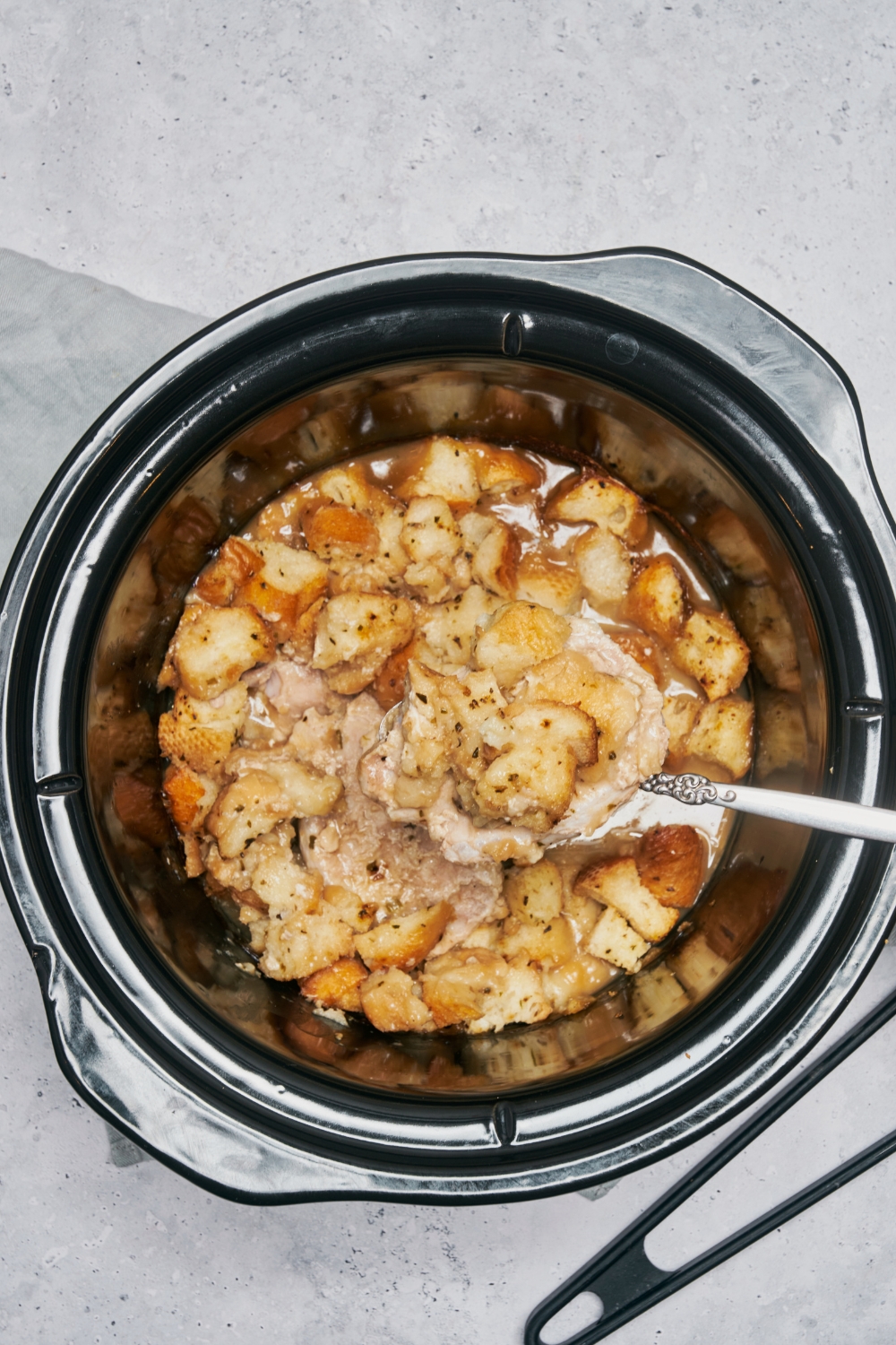 A slow cooker filled with stuffing in a creamy soup mixture and a large spoon is scooping out the stuffing.