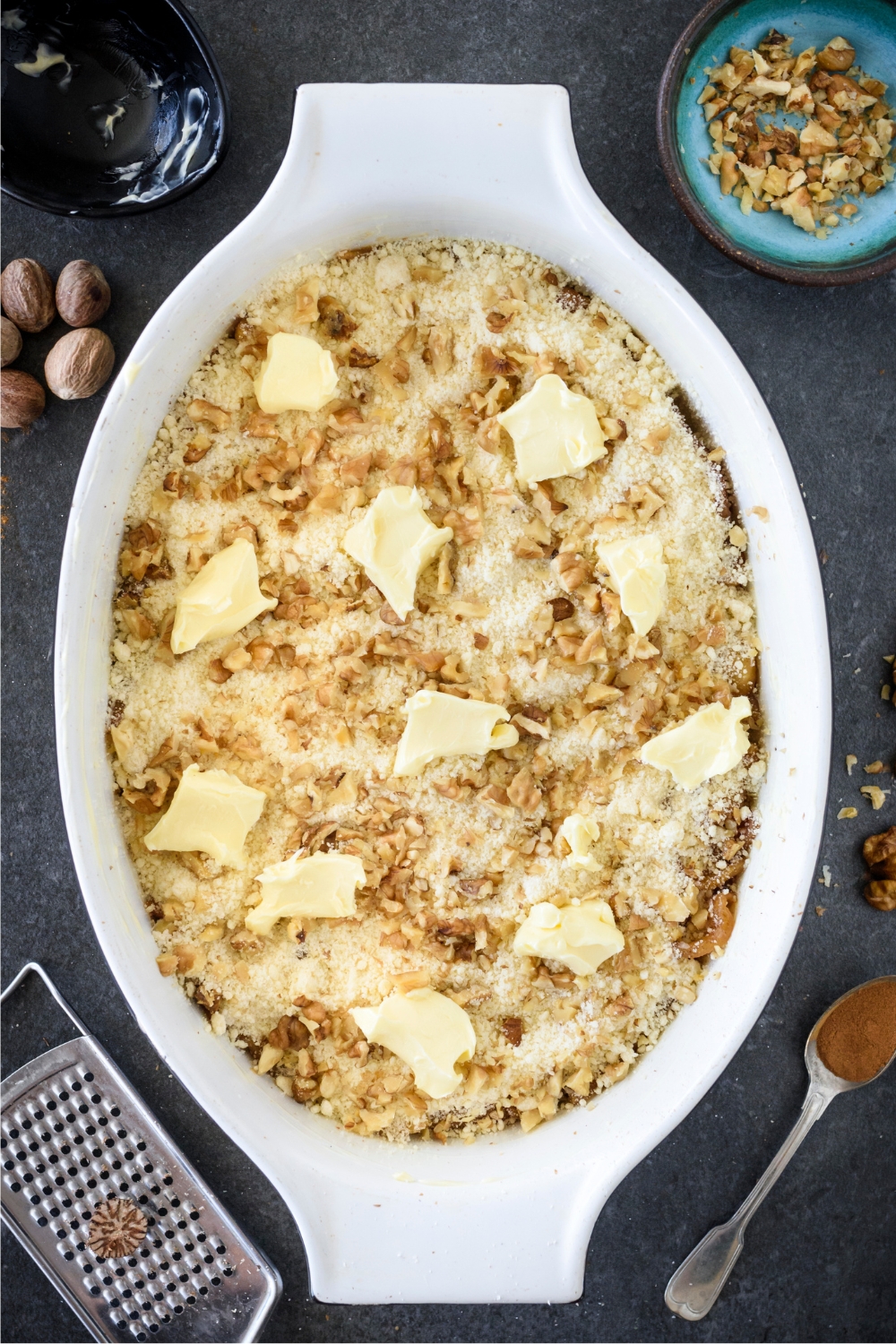 A baking dish filled with unbaked apple dump cake covered in a layer of cake mix, chopped walnuts, and chunks of unmelted butter.